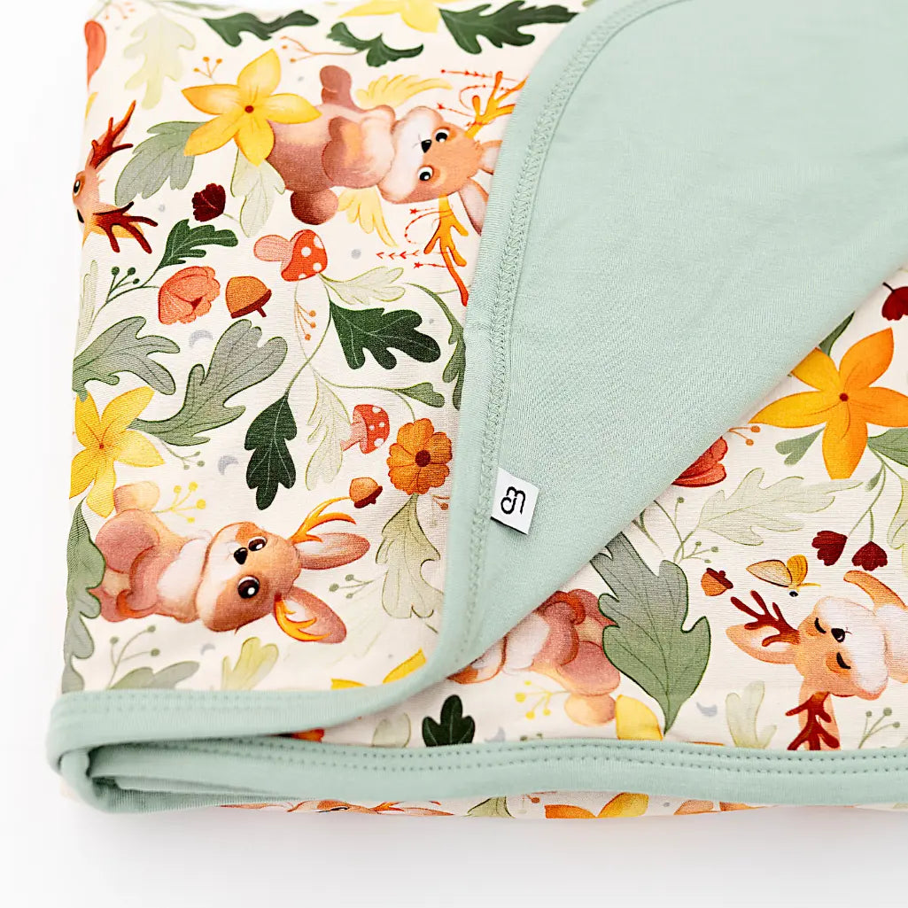 A close up of a folded blanket with a sage green reverse and hem, printed with antlered bunnies, mushrooms, and yellow flowers.